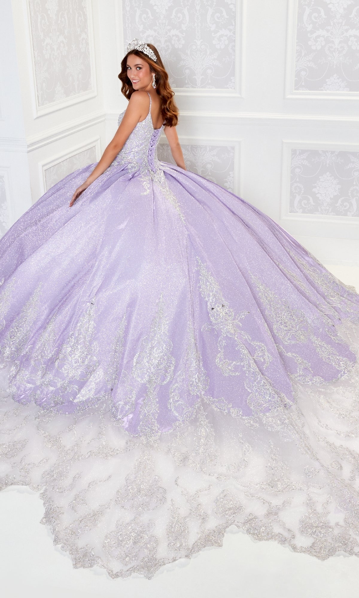 A woman wearing the Quinceanera Dress PR22141 By Princesa, a sparkling lavender ball gown with silver embroidery and a corset back, stands in an ornate white room, looking over her shoulder while wearing a tiara.